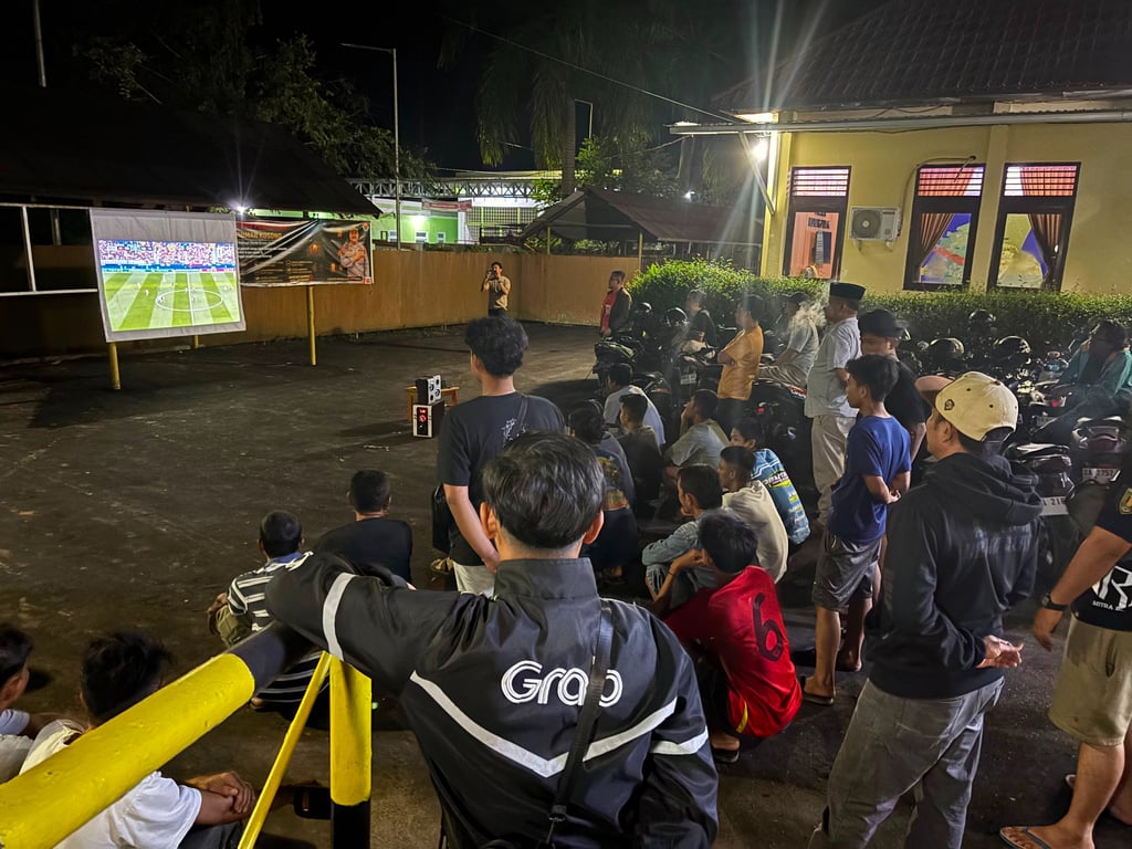 A crowd of people watching a football match on a large projector screen outdoors at night.