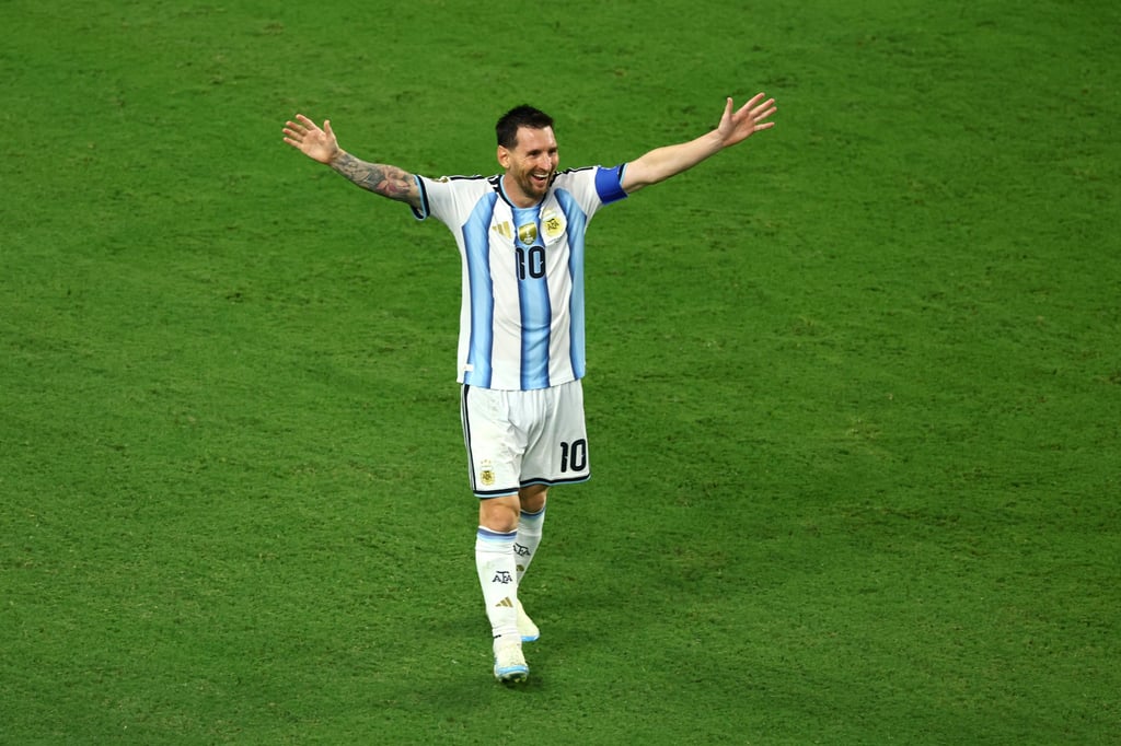 Lionel Messi celebrating in an Argentina jersey with arms wide on a green soccer pitch.
