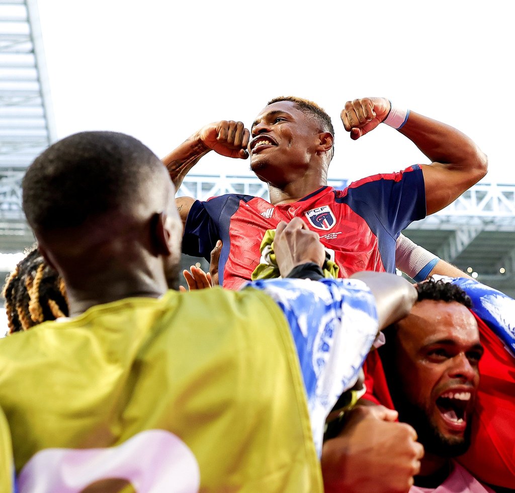 A smiling soccer player in a blue and red jersey celebrates a goal with teammates in a stadium.