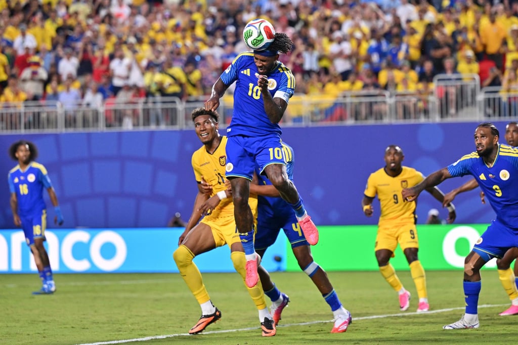 A professional soccer player in a blue jersey jumps high to head the ball during a competitive match.