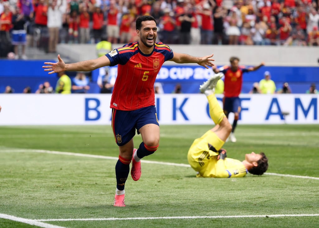 Spanish soccer player celebrates a goal on the field while the opposing goalkeeper lies on the grass.