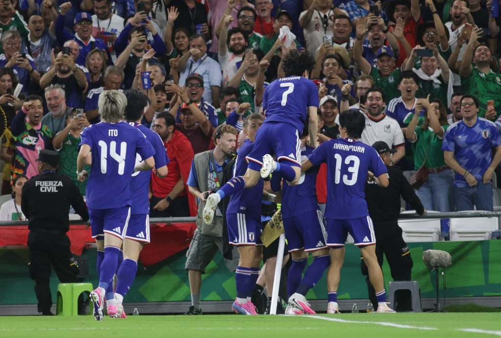 Japan national football team players in blue jerseys celebrate a goal in front of cheering fans.