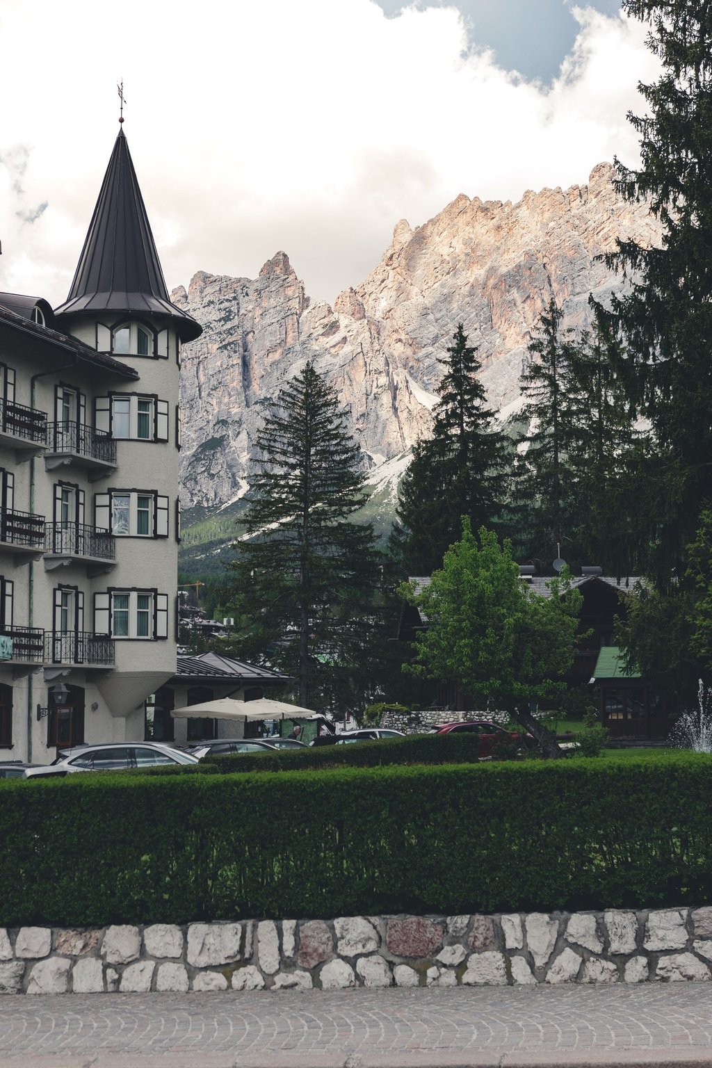 A classic alpine hotel with a turret overlooks pine trees and the jagged peaks of the Dolomites mountains.