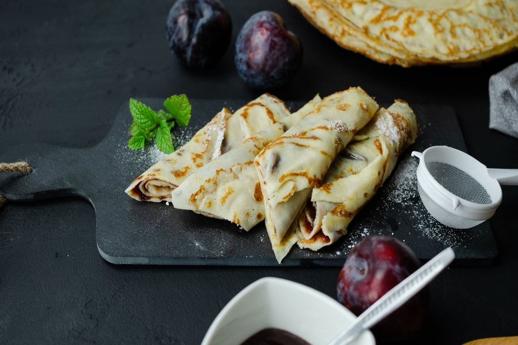 Rolled crepes with plum jam filling on a black slate board with fresh mint and powdered sugar.