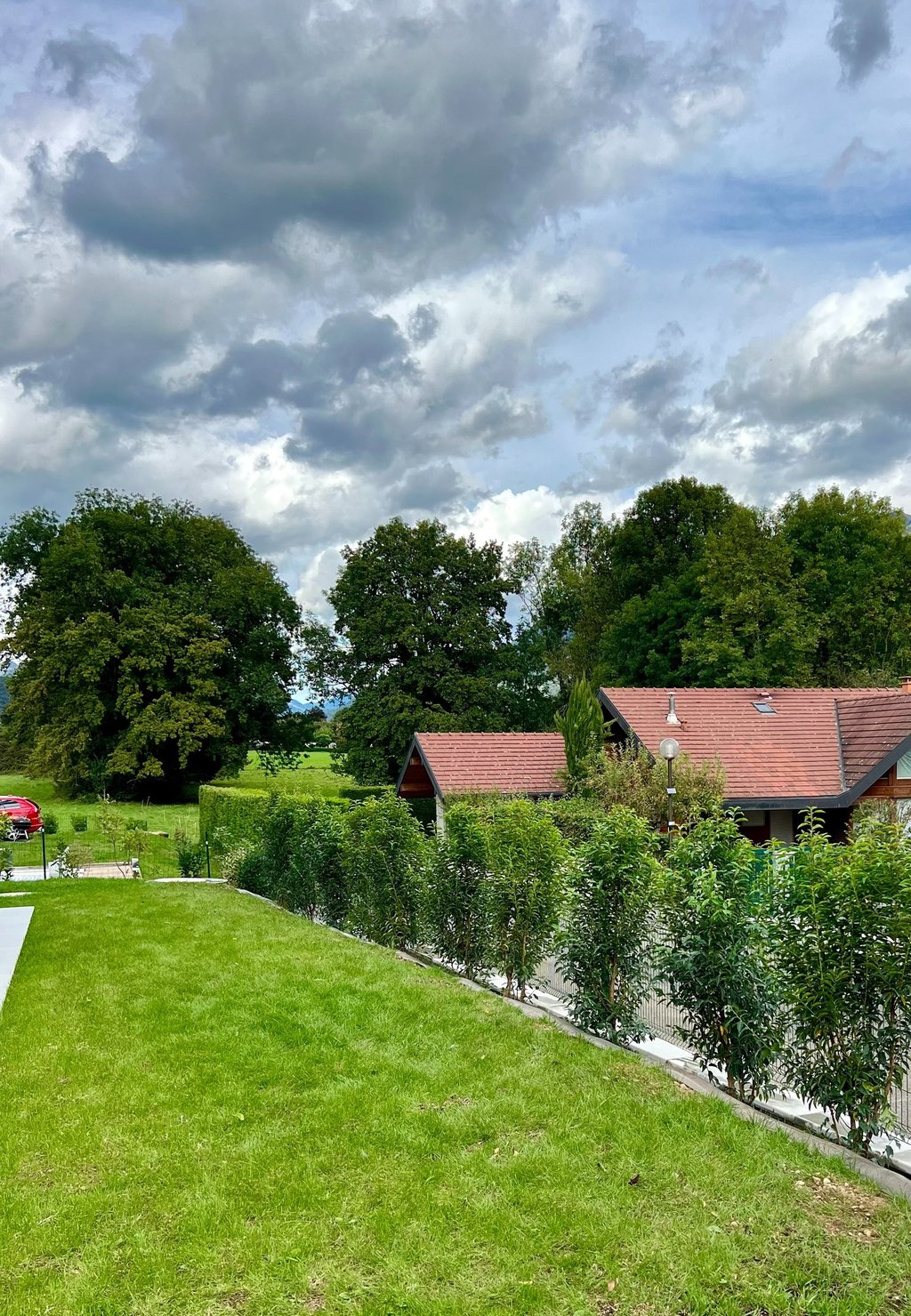 Lush green lawn leading to a modern house with a red tile roof, surrounded by mature trees under a cloudy sky.