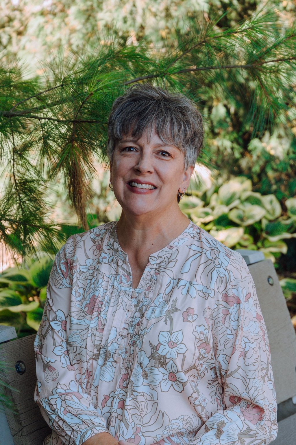Smiling woman with short gray hair wearing a floral print blouse while sitting outdoors in a garden.
