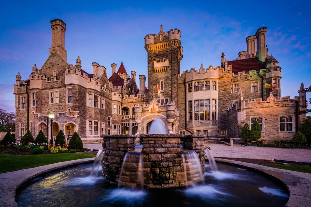 Illuminated stone fountain in front of Casa Loma Gothic Revival castle in Toronto at dusk.