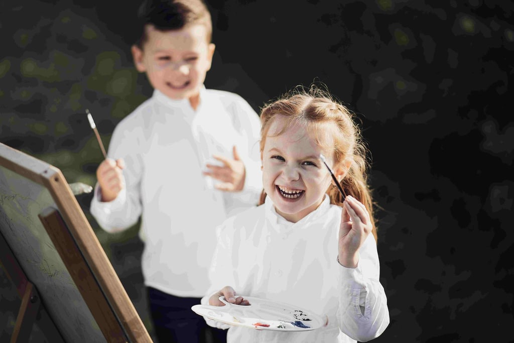 Two kids, wearing white shirts, happily paint outdoors.