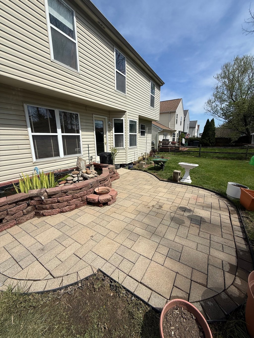 A residential backyard featuring a tan stone paver patio, stone retaining wall, and beige vinyl siding house.