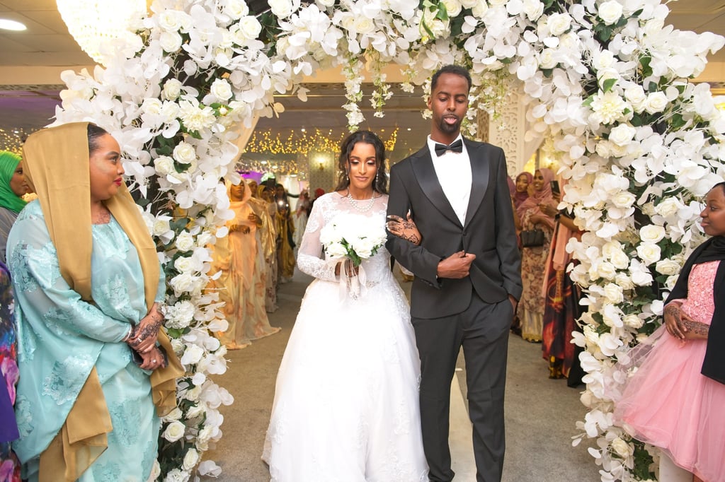 bride in white wedding dress linking arms with groom in black suit, entering the venue, floral arch framing them
