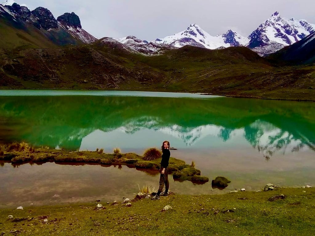 A hiker stands by a turquoise alpine lake reflecting snow-capped Andes mountains in Peru.