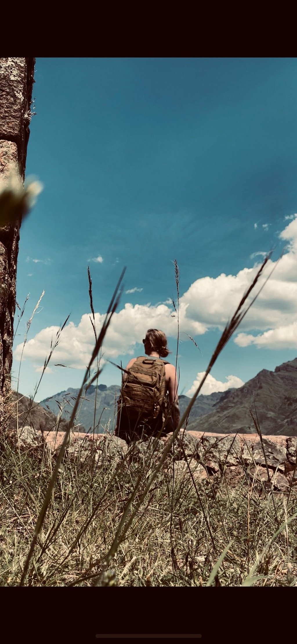 A hiker with a backpack sits on a ridge overlooking a vast mountain range under a blue sky.