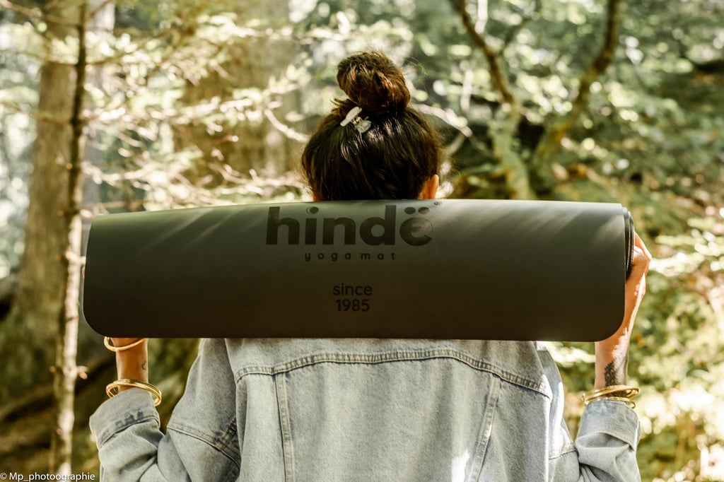 A woman in a denim jacket carrying a black Hindë yoga mat on her shoulders in a sunny forest.