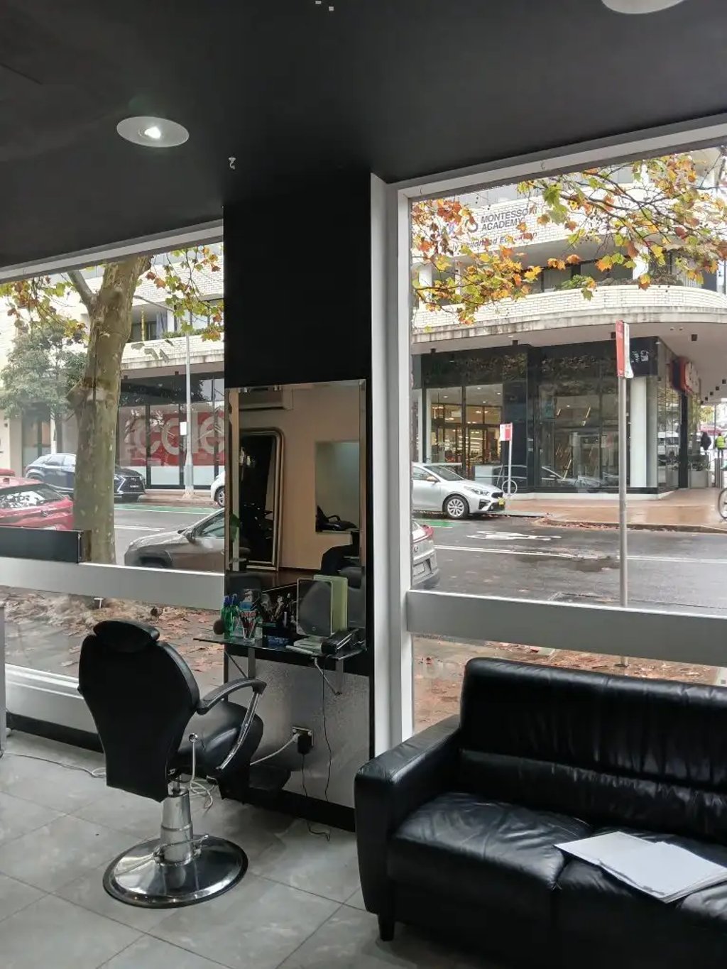 Modern barbershop interior featuring a black leather salon chair, mirror, and waiting sofa near a large window.