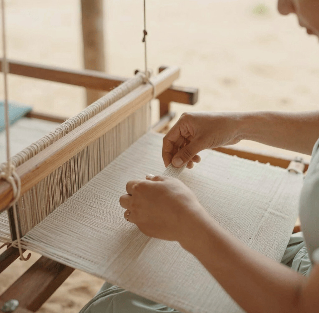 An intimate close-up of a local artisan hand-weaving textiles on a traditional wooden loom, soft side light, warm sand tones.