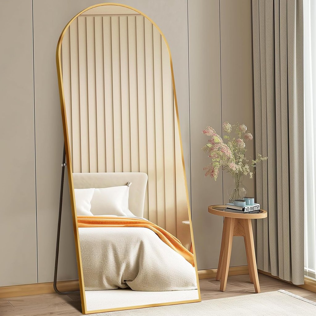Full length arched floor mirror with gold frame leaning against a bedroom wall next to a wooden side table.