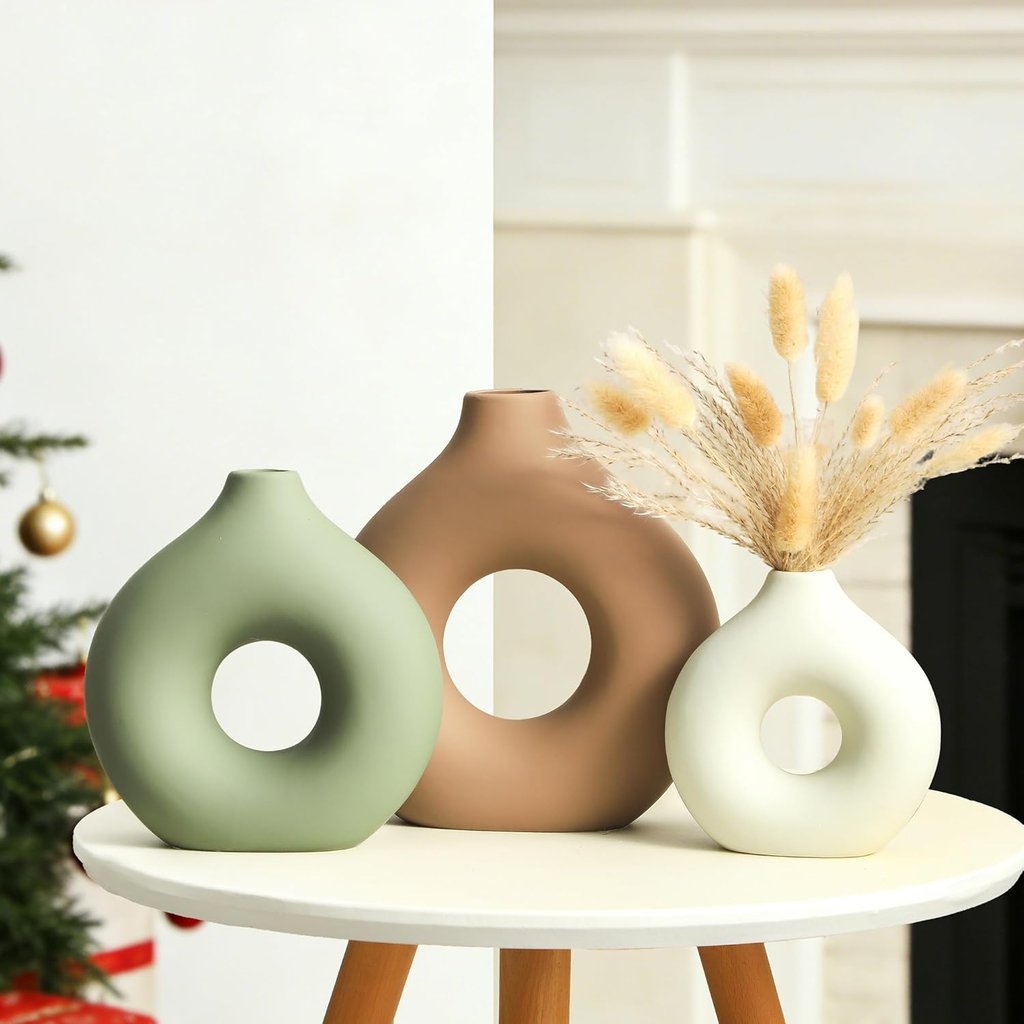Set of three modern ceramic donut vases in sage green, tan, and white on a side table.