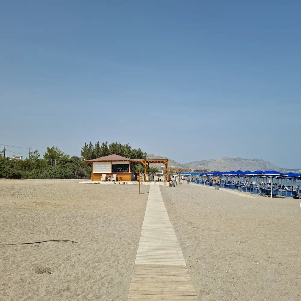 A wooden boardwalk leads to a beach bar with blue umbrellas on a sunny sandy shore.