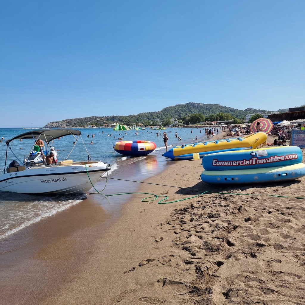 A motorboat and inflatable water sports towables sit on a crowded sandy beach under a clear blue sky.
