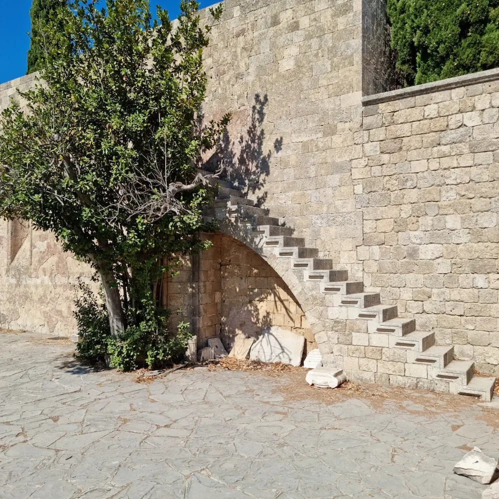 Ancient stone steps built into a medieval fortress wall next to a green tree in Rhodes, Greece.