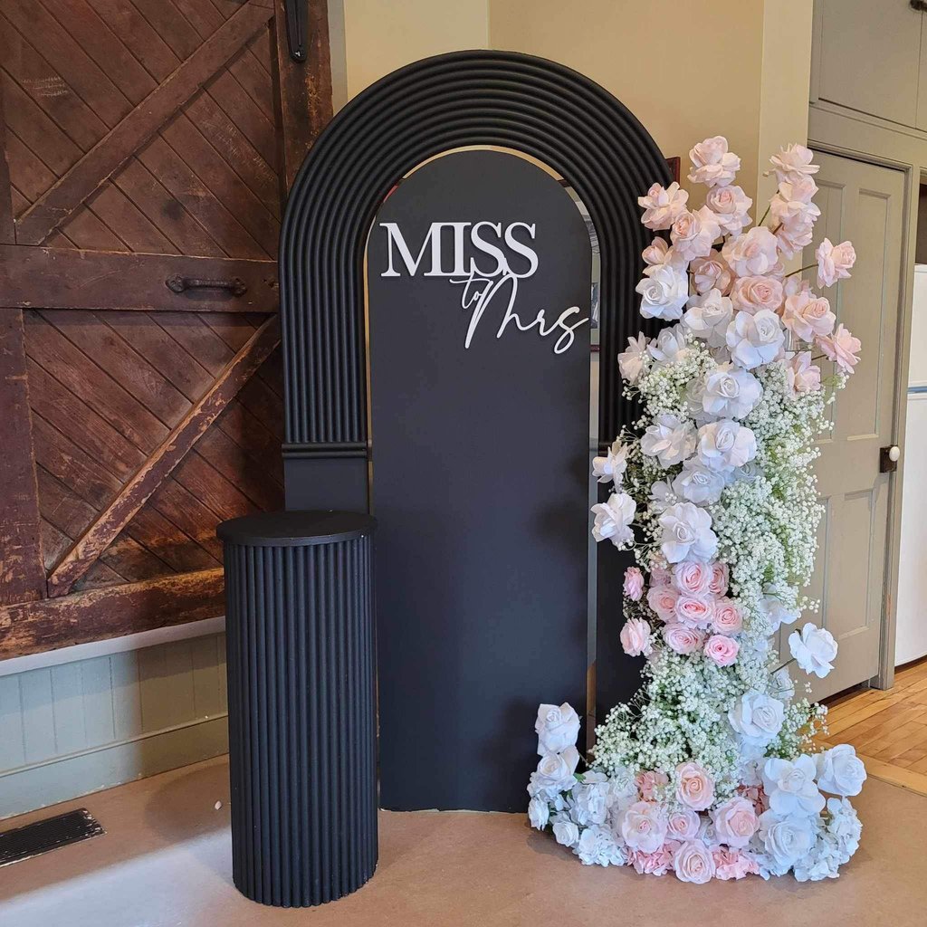 Black backdrop with a white Miss To Mrs sign and pink, white florals with baby breaths