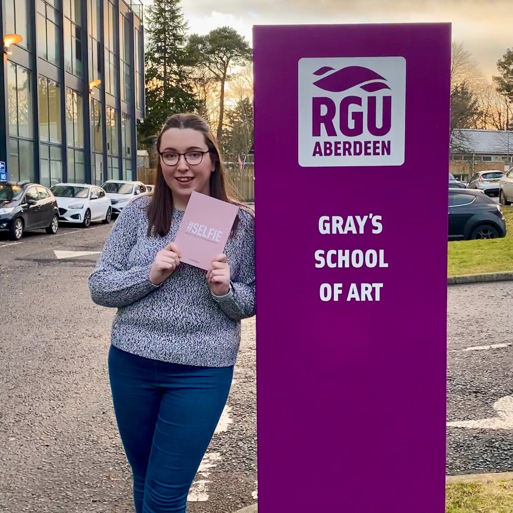 Erin, the owner of EB Socials, standing beside a sign for 'Gray's School of Art'