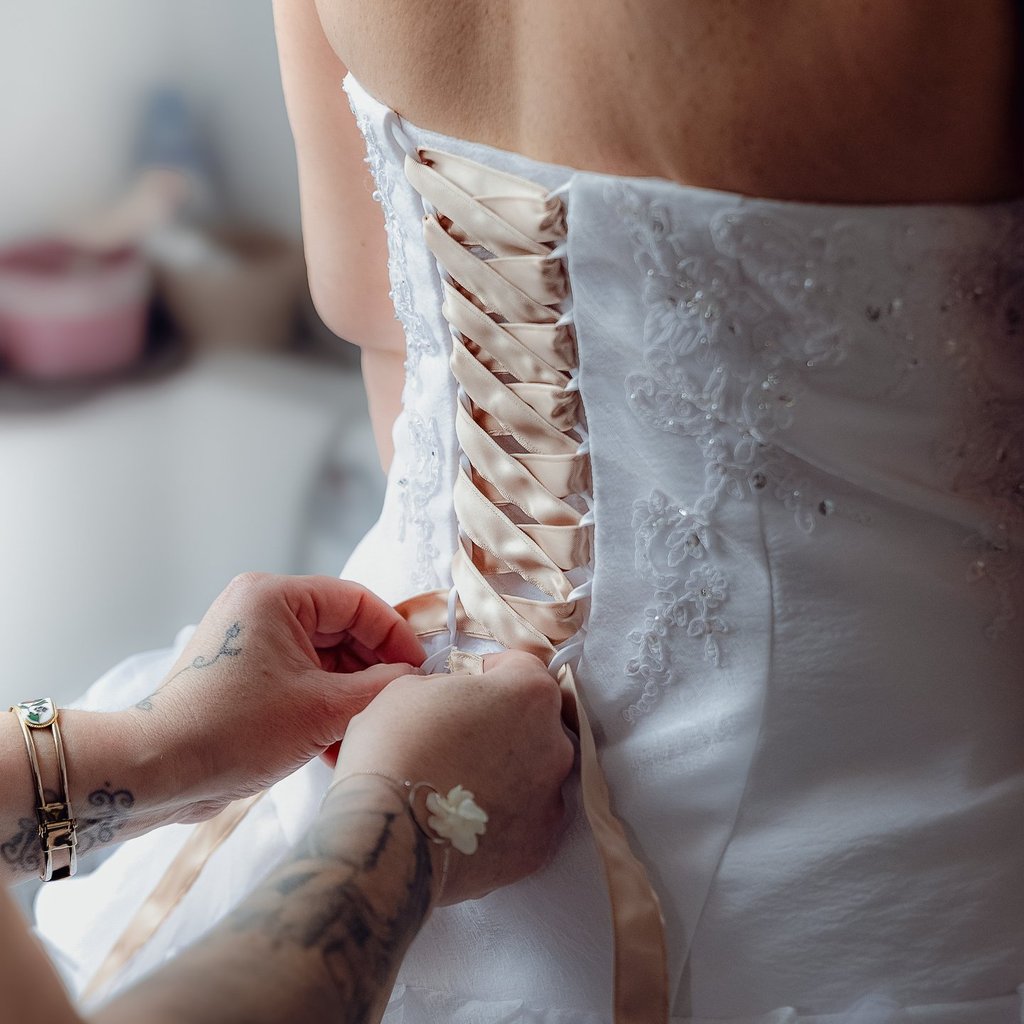 Gros plan sur une mariée se faisant lacer sa robe de mariée en dentelle blanche avec un ruban de cor