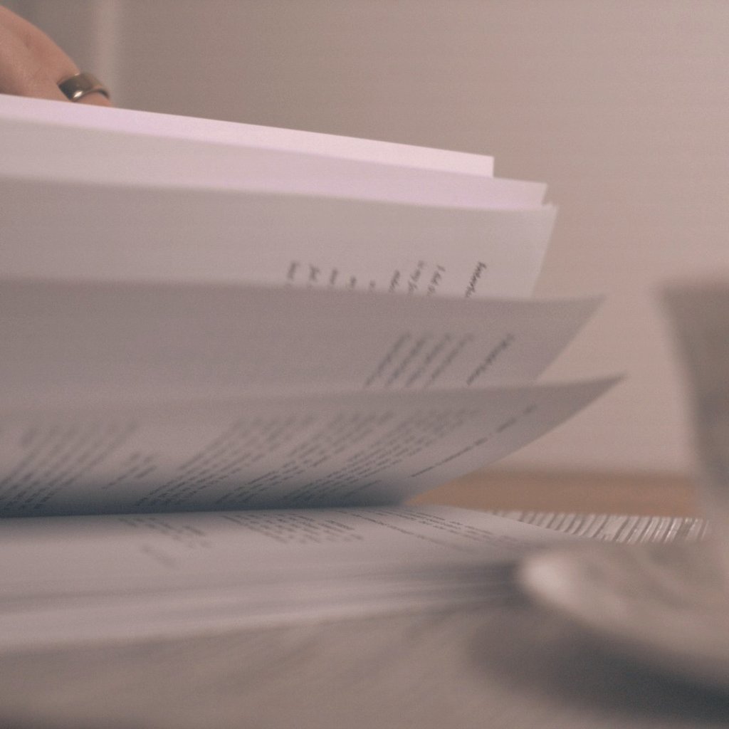 Close-up of a person's hand flipping through book pages next to a coffee cup on a table.