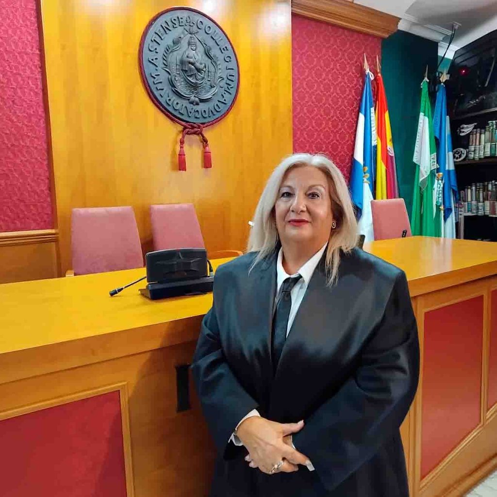 Abogada con bata negra de pie en una sala del tribunal con banderas oficiales y un emblema judicial