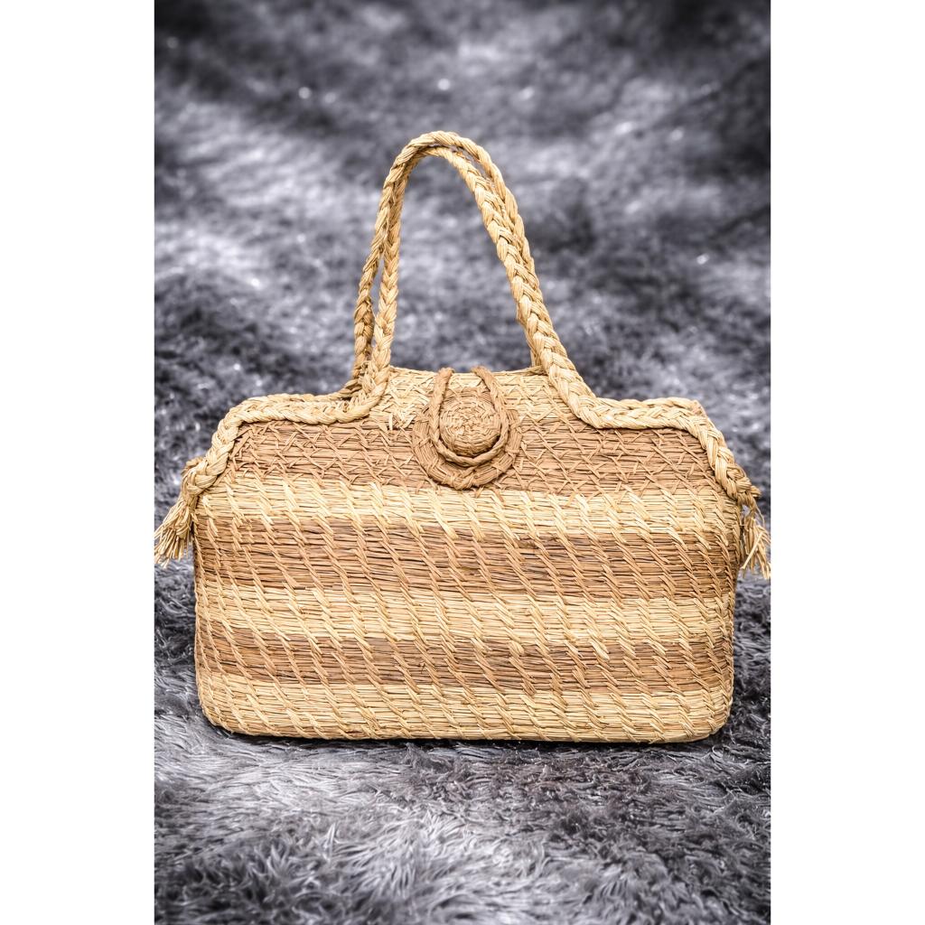 Handwoven straw tote bag with braided handles and horizontal striped texture on a grey faux fur background.