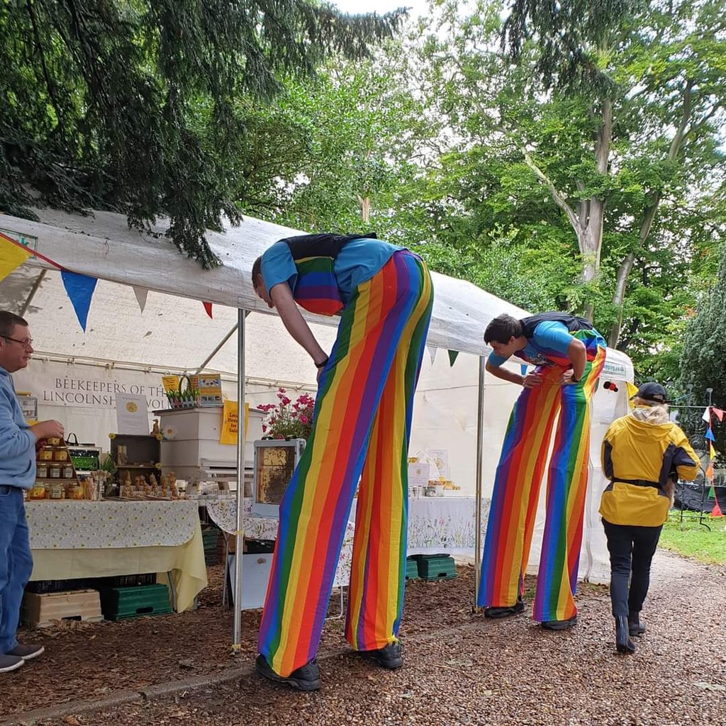 Two stilt walkers in rainbow striped pants visit a honey vendor stall at an outdoor festival.