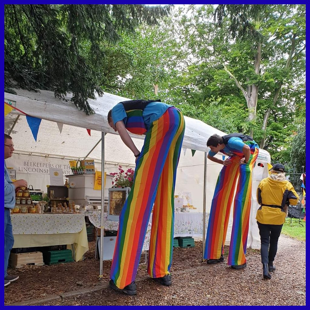 Performers on stilts wearing rainbow trousers at a beekeeping festival market stall outdoors.