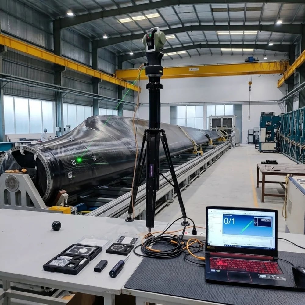 Precision laser tracker measuring a large composite wind turbine blade in an industrial manufacturing facility.
