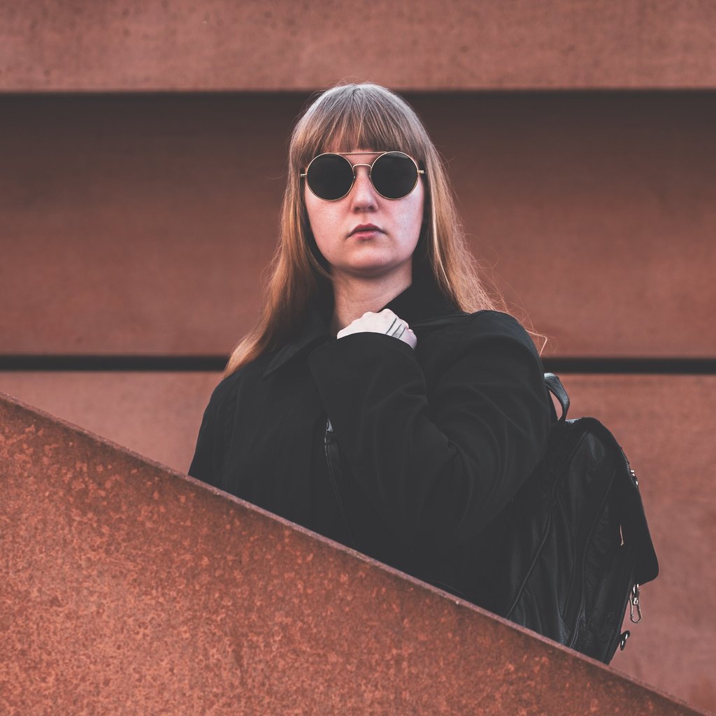 a woman in sunglasses and a black jacket standing in between a wall and a staircase