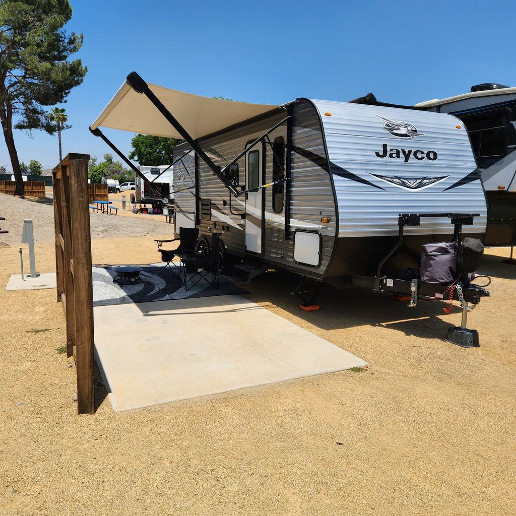 A Jayco travel trailer parked at a gravel RV campsite with the awning extended over a concrete patio.