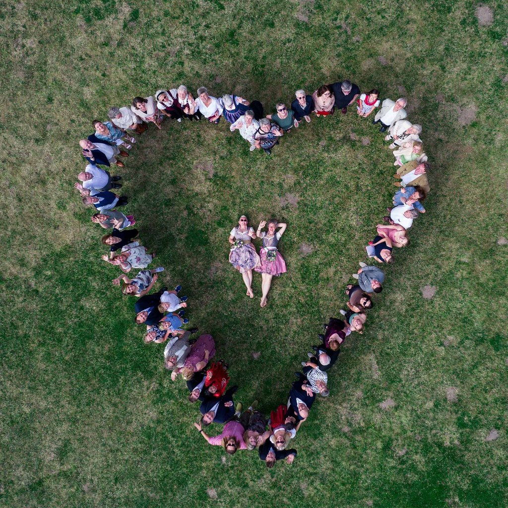 Hochzeitsgesellschaft formt Herz auf Wiese um liegendes Brautpaar. Drohnenaufnahme von Joel Pingel.