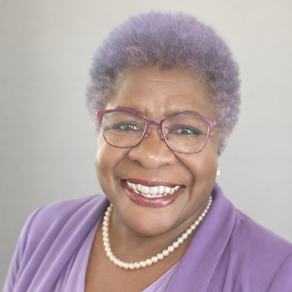 Smiling woman with short purple hair wearing glasses, a pearl necklace, and a purple blazer.