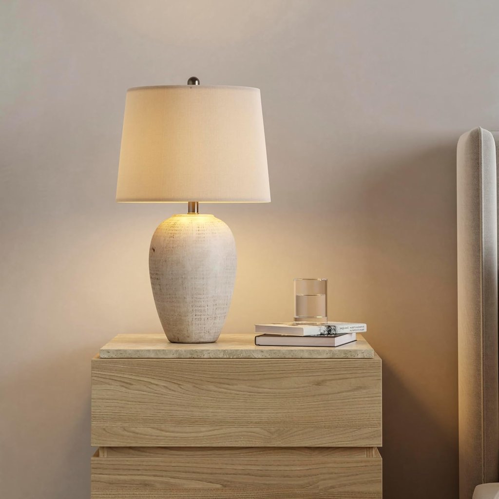 Ceramic table lamp with textured base on a light wood nightstand in a neutral bedroom.
