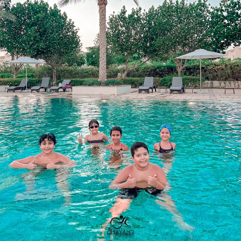 A group of happy children smiling and swimming in a resort swimming pool with lounge chairs and palm trees.