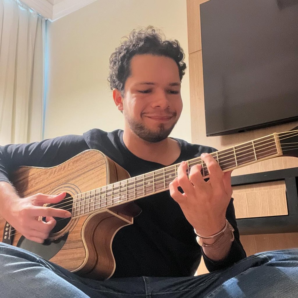 Músico e palestrante autista Lucas Sampaio, com violão marrom em quarto de hotel.