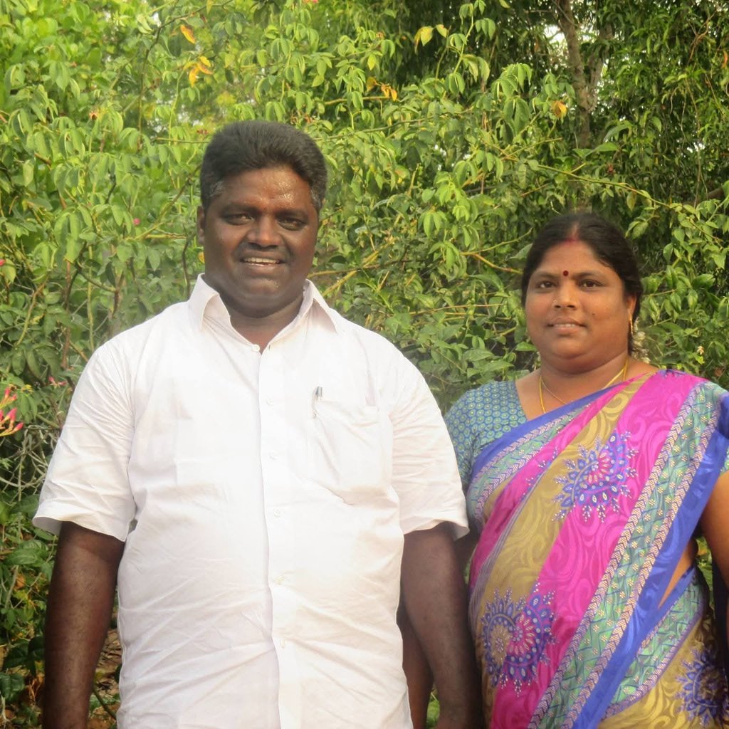 A smiling Indian couple posing together outdoors against a lush green leafy background.