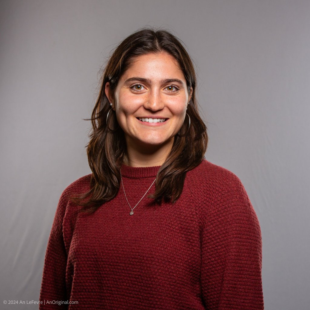 Professional headshot of a smiling woman with brown hair wearing a maroon sweater against a grey background.