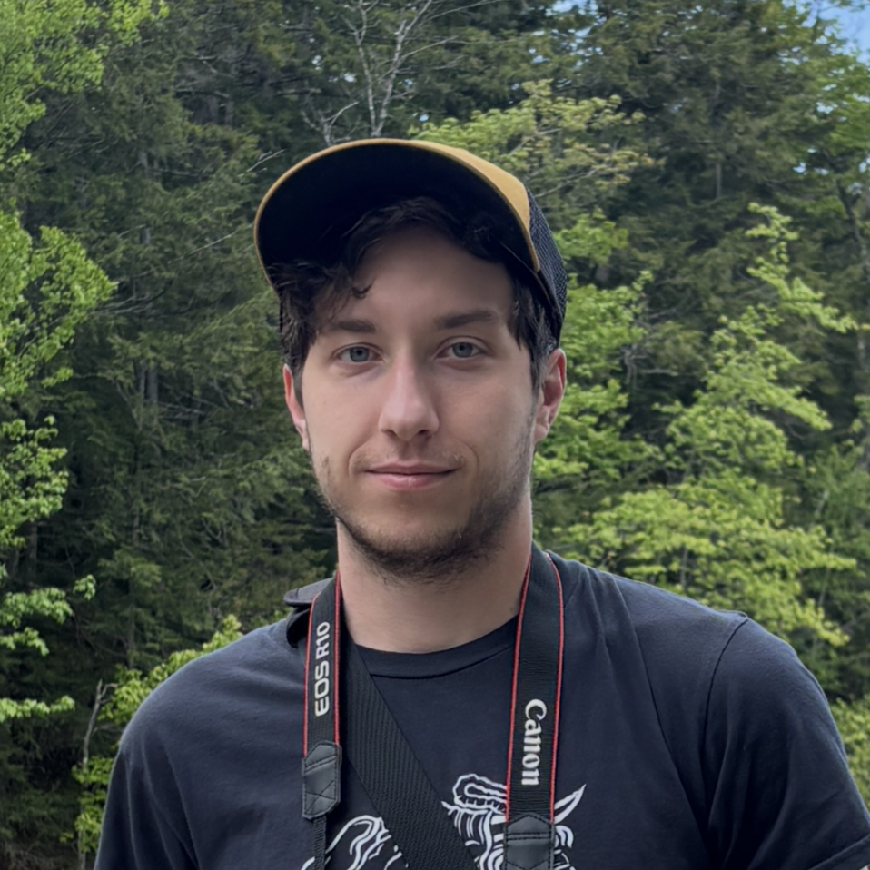 Young male photographer wearing a yellow hat and Canon EOS R10 camera outdoors with forest background.