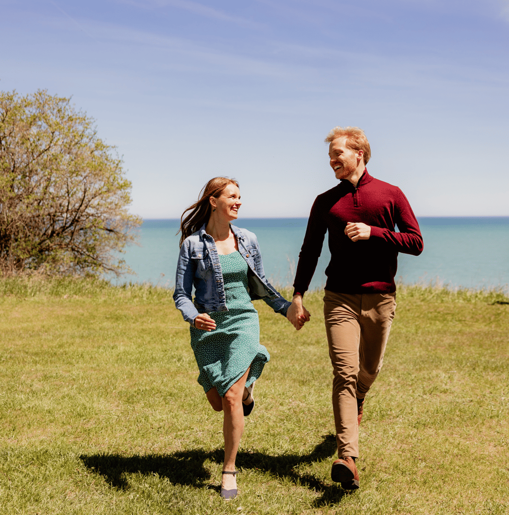 Happy couple holding hands and running through a grassy field overlooking the blue ocean on a sunny day.