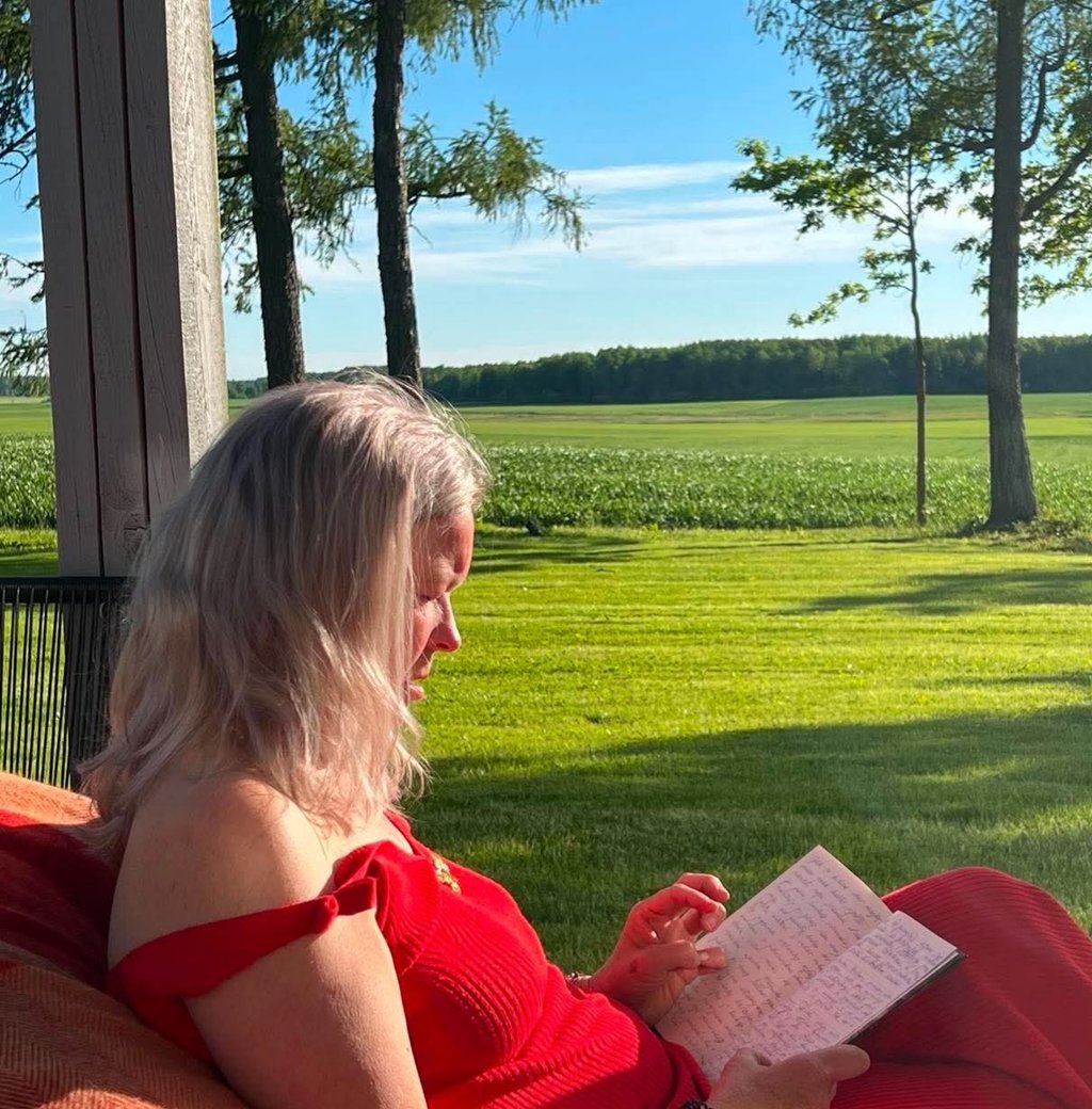 A woman in a red dress sits on a porch reading a book overlooking a green field and forest.