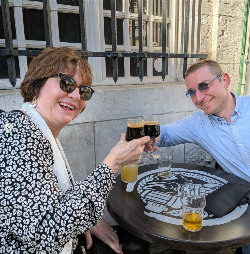 A smiling man and woman toast with dark craft beers at an outdoor pub table.