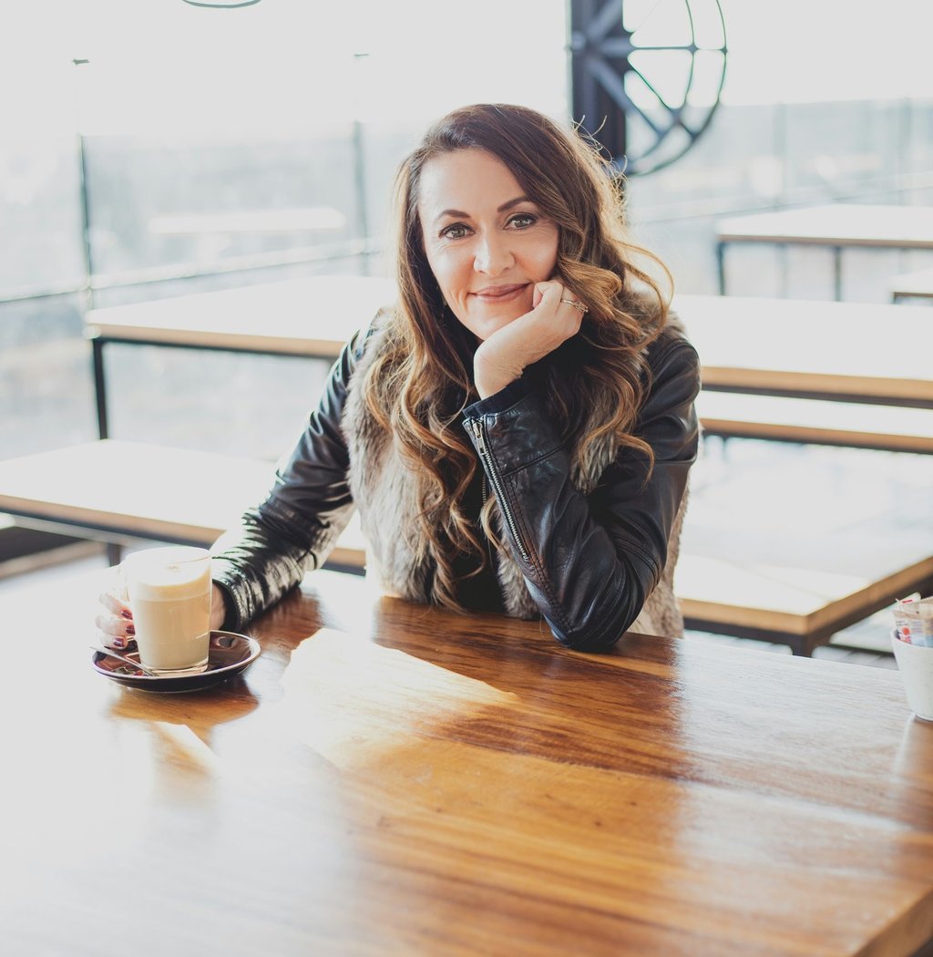 Smiling woman in a black leather jacket enjoying a latte coffee at a bright modern cafe table.