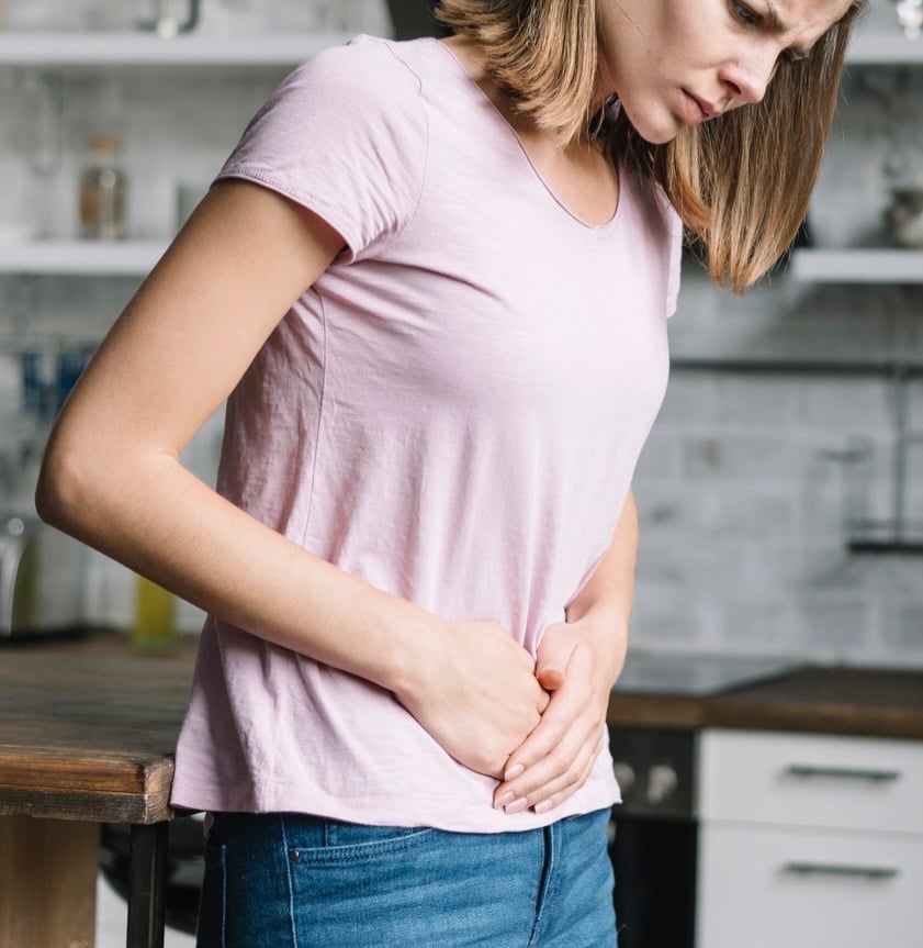 A young woman holding her stomach in pain due to digestive issues or menstrual cramps in a kitchen.