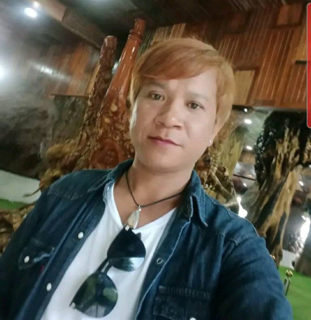 Man smiling indoors in Pattaya, Thailand, wearing a denim shirt and sunglasses on a white T-shirt.