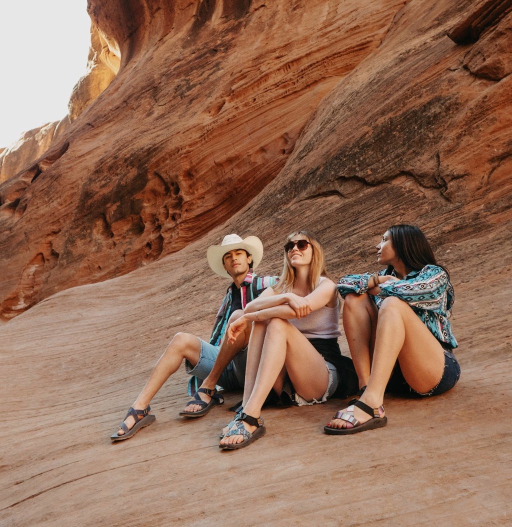 Friends sitting on red rock canyon formations wearing casual summer hiking clothes and outdoor sandals.